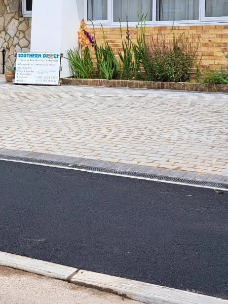 A block paved driveway in front of a house with a brick wall and window. A sign on the left reads "SOUTHERN DRIVES" and provides contact information. The block paving area is bordered by an asphalt road in the foreground and plants to the right.