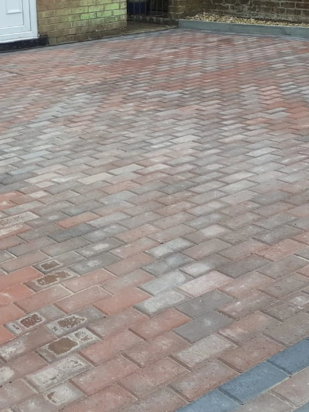 A driveway with a herringbone pattern made of block paving in various shades of red and gray. The edge of the driveway is bordered by darker gray bricks, and a partially visible white door is seen in the background.