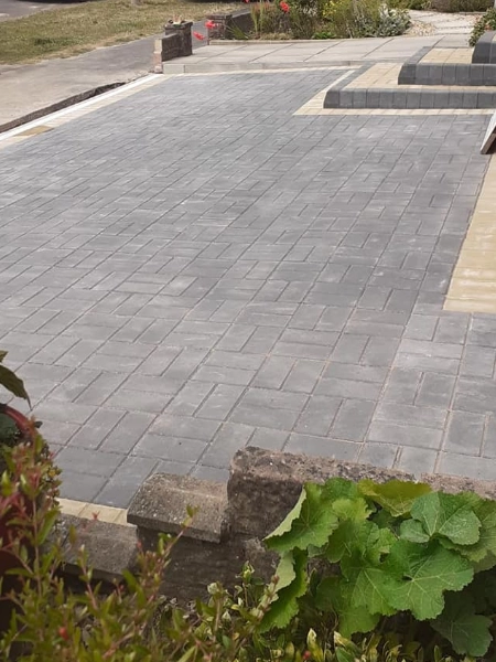 A newly paved driveway with gray blocks leads to a raised area with stairs. Green plants and shrubs adorn the foreground, while the background showcases a residential street with more greenery.