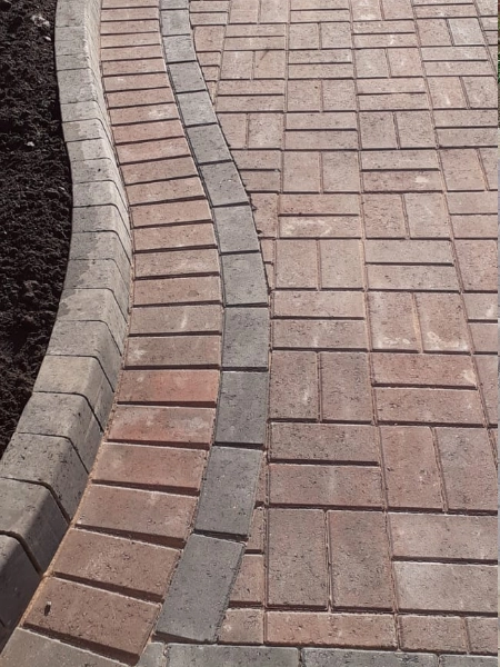 A path made of block paving in varying shades of red and gray forms a curved design. The border of the path is composed of darker gray bricks. 