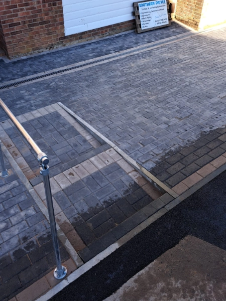 A newly paved driveway in front of a brick building features elegant block paving with interlocking grey bricks. A sturdy metal handrail graces the left side, and a Southern Drives sign is visible on the building in the background.