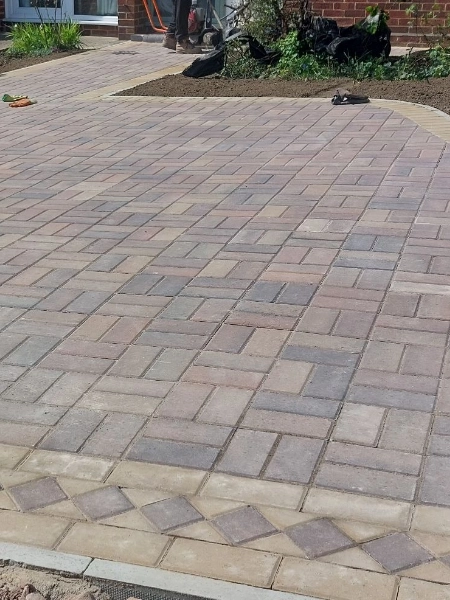 A neatly laid driveway with a light and dark block paving pattern, bordered with a contrasting diamond-shaped design. In the background, there is a garden bed, some plants, and construction tools.