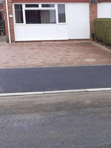 A newly-paved driveway in front of a house with a brick exterior and white front facade features light brown pavers, bordered by a hedge on one side. Dropped kerbs provide easy access from the road in front of the property.