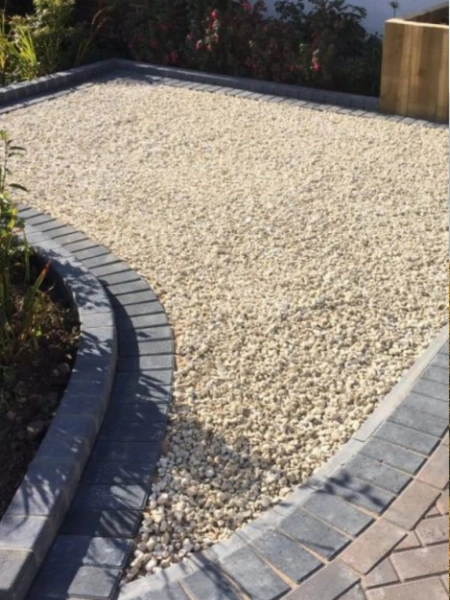 A rectangular garden bed is bordered by dark, grey bricks and filled with small, light-colored pebbles. A curved pathway made of the same grey bricks runs along the edge. Flowering bushes with green leaves are visible in the background, complementing the natural stone aesthetic.