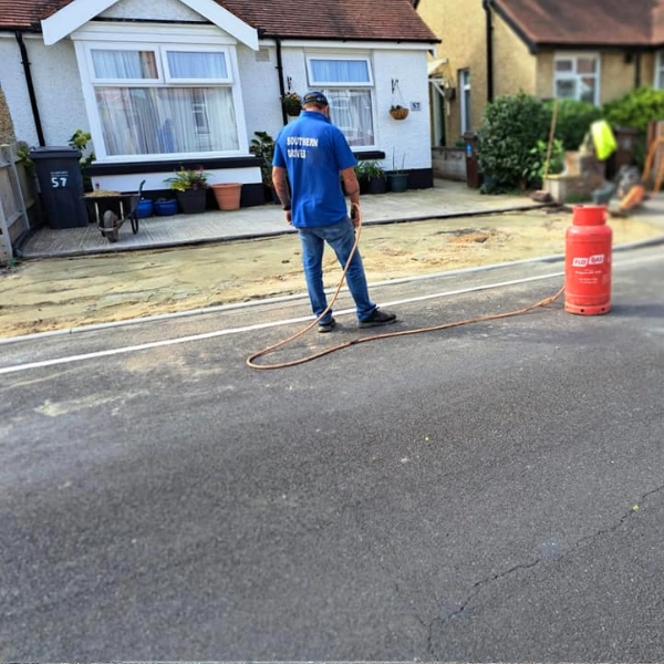A southern drives worker in a blue uniform operates a gas-powered line-painting machine to mark street lines in front of residential houses, an essential part of dropped kerb services. A large red gas cylinder is placed beside them on the road's edge. Bushes and potted plants are visible in front of the houses.