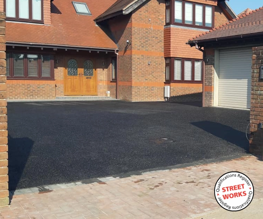 A new tarmac driveway by Southern Drives. The house features gable roofs and a garage door on the right. A round sticker with "Street Works" and related qualifications is at the bottom right corner of the image.